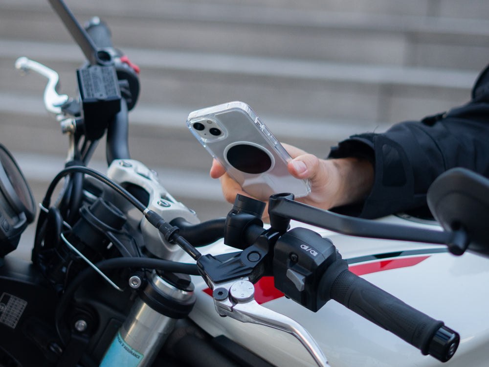 Motorista fijando su smartphone en el soporte de teléfono moto pro con placa montado en el manillar de su moto. El soporte magnético garantiza una fijación segura y rápida, ideal para la navegación y el uso del teléfono con total seguridad en la carretera.